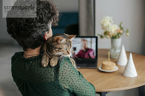 Woman with cat on shoulders celebrating birthday through video call at home