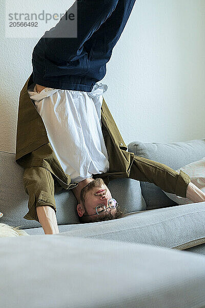 Young active man doing headstand on sofa at home