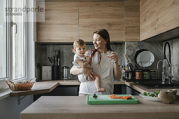 Smiling woman with glass of water carrying daughter in kitchen