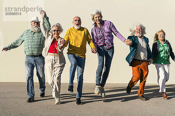 Excited retired men and women holding hands walking in front of wall on sunny day