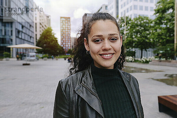 Smiling beautiful young woman wearing black leather jacket standing in city