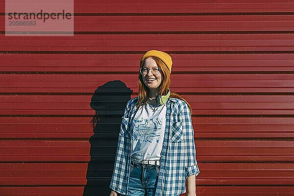 Smiling teenage girl in yellow cap and glasses standing in front of corrugated iron