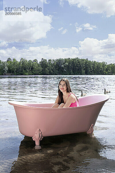 Happy girl sitting inside pink bathtub under cloudy sky in lake