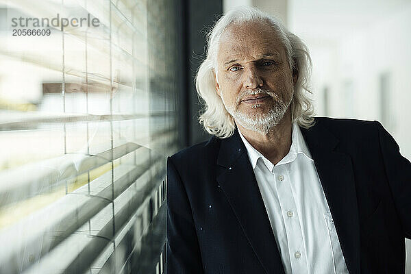 Confident senior businessman leaning on window glass in office