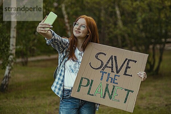 Smiling girl holding protest banner and taking selfie through smart phone at public park