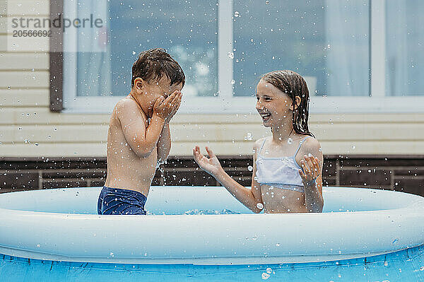 Happy sibling splashing water in inflatable swimming pool at back yard