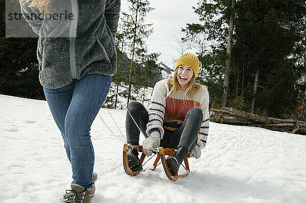 Friend pulling cheerful woman sitting on sled in winter vacation