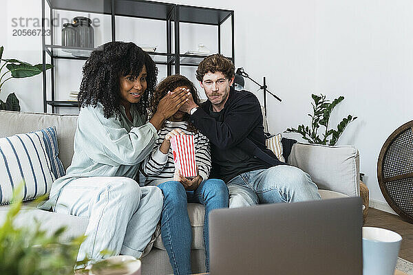 Daughter sitting on sofa watching movie with parents covering her eyes to protect her