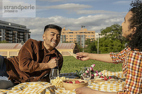 Girlfriend giving snack to boyfriend lying on picnic blanket at park