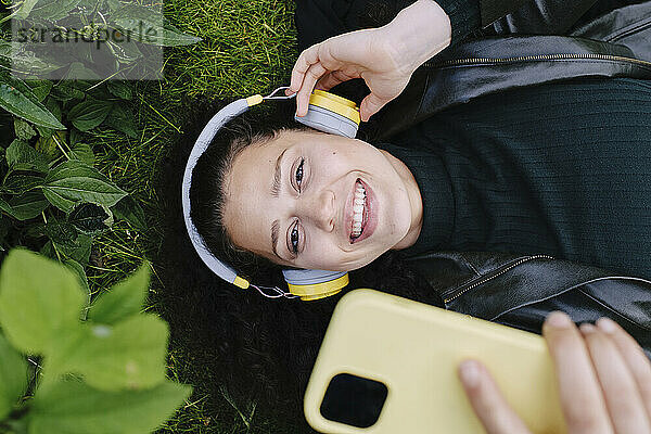 Happy beautiful woman using smart phone and listening to music through wireless headphones lying on grass at park