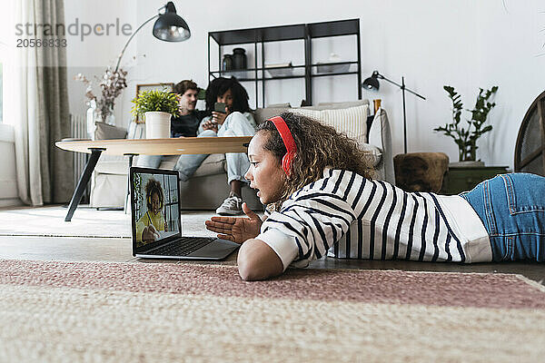 Girl having video call with friend lying on floor iwth parents sitting on couch in background