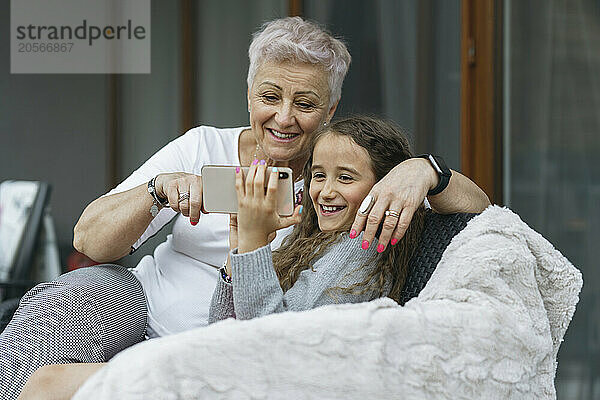 Happy grandmother using mobile phone with cute granddaughter at patio