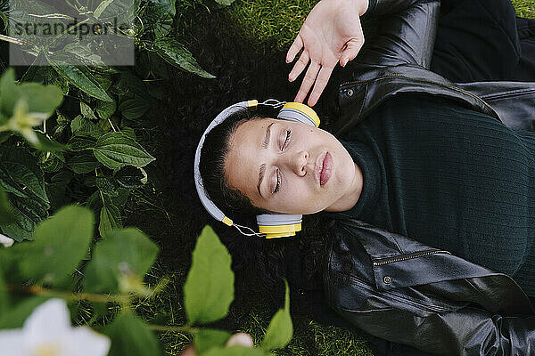 Young woman with eyes closed listening to music through wireless headphones lying near green plants at park