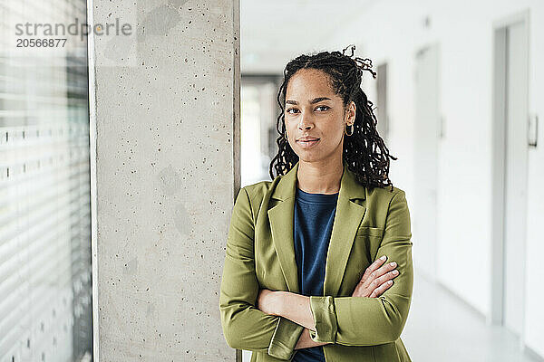 Confident young businesswoman with arms crossed leaning on column in office corridor