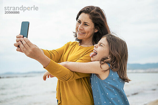 Happy woman taking selfie through smart phone with daughter at beach