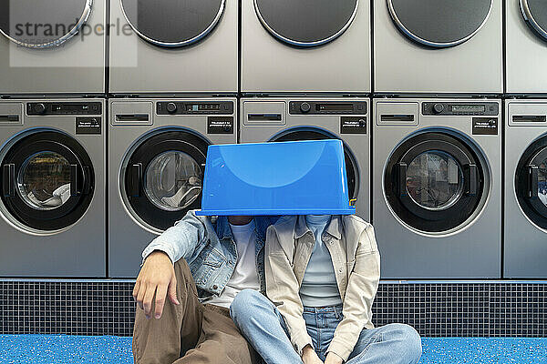 Playful young couple with faces covered in blue basket at laundromat