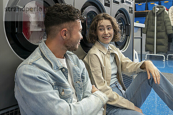 Smiling young friends leaning on washing machines sitting at laundromat