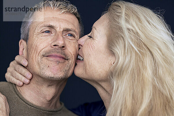 Smiling blond senior woman kissing man on cheek against black background