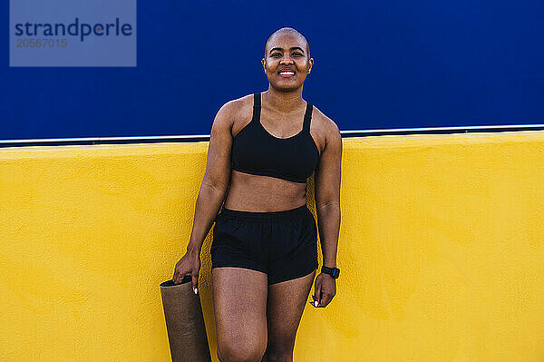 Smiling bald woman holding mat and standing in front of wall