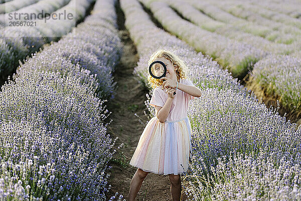 Playful girl holding magnifying glass and standing in lavender field