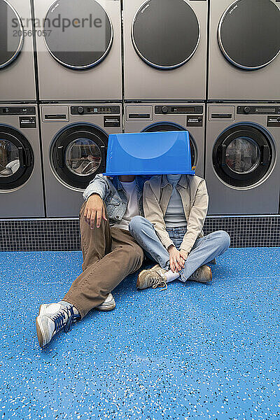 Playful young boyfriend and girlfriend with faces covered in blue basket at laundromat