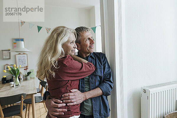 Smiling senior woman and man hugging and leaning on white wall at home