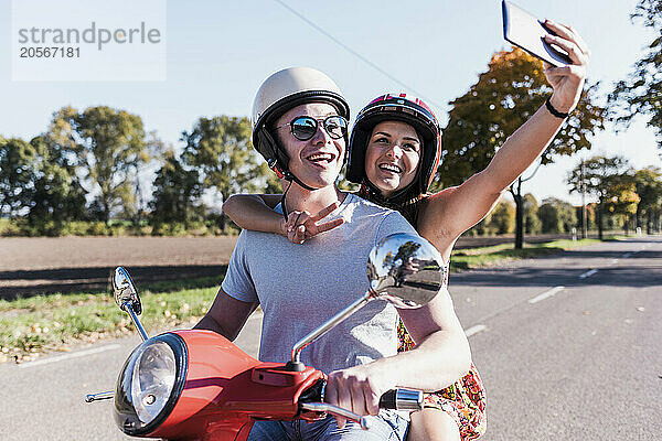 Happy young woman making peace sign and taking selfie with boyfriend on motor scooter at country road