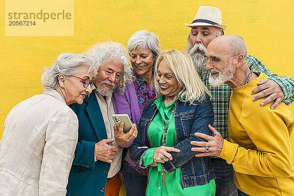 Cheerful elderly woman sharing smart phone with friends in front of wall