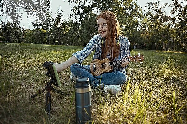 Redhead girl holding ukulele and vlogging through smart phone at public park