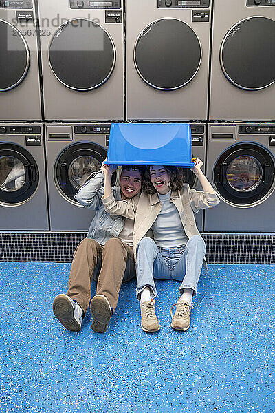 Cheerful young couple with blue basket over head sitting in front of washing machines at laundromat