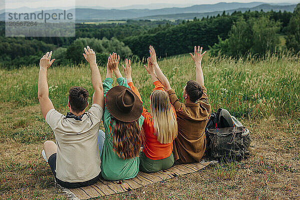 Group of young couples raising hands and sitting on meadow at mountain