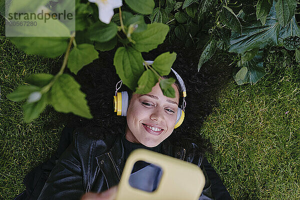 Smiling young woman using smart phone and listening to music through wireless headphones lying on grass at park