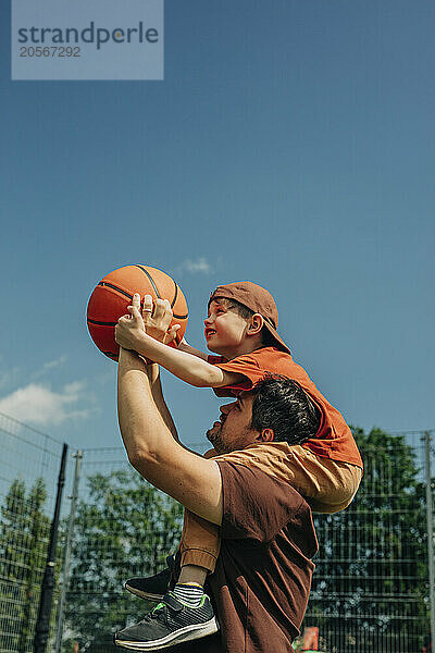 Father carrying son on shoulder and throwing basketball at court