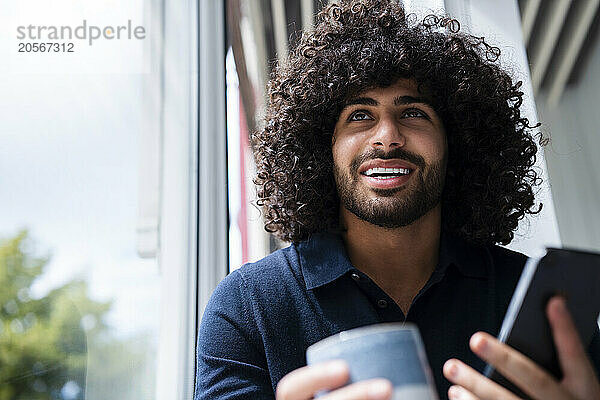 Smiling young businessman with coffee cup and smart phone by window at workplace