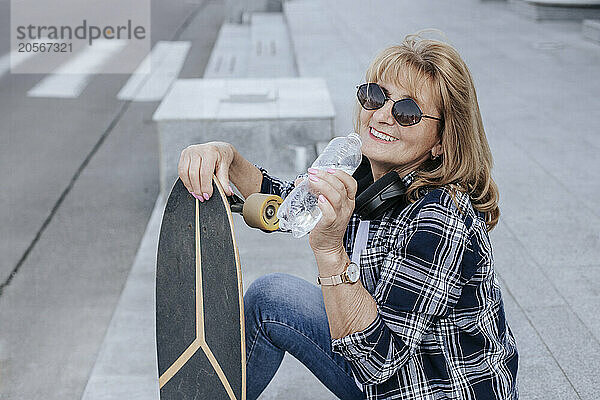 Smiling senior woman drinking water and sitting with skateboard on stairs at park