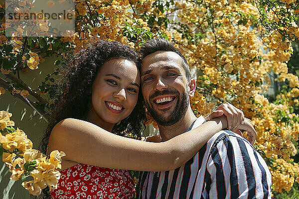 Happy woman with arms around boyfriend amidst yellow bougainvillea vine