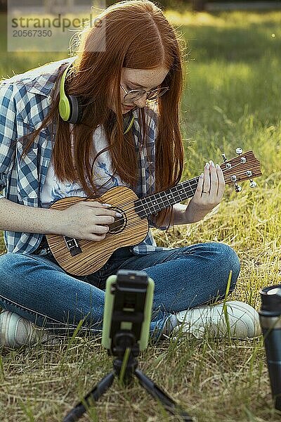 Redhead girl playing ukulele and sitting cross-legged at public park