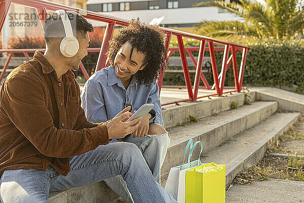 Smiling woman sitting on staircase with boyfriend wearing wireless headphones at park