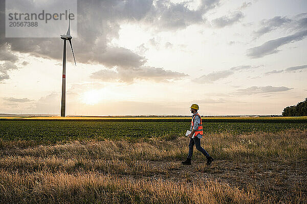 Engineer walking in field with wind turbine at sunset