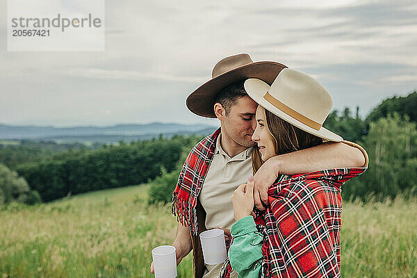 Affectionate young man with arm around girlfriend wearing plaid shawl on meadow in mountain