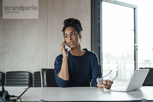 Young businesswoman with eyeglasses and laptop talking on smart phone at desk in office