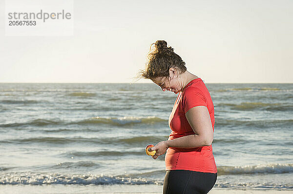 Cheerful woman holding apple and laughing near sea at beach