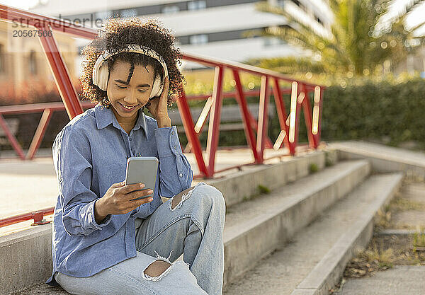 Smiling woman wearing wireless headphones and using smart phone sitting on staircase at park