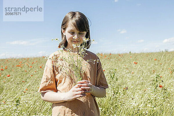 Smiling girl holding flowers and standing in meadow under blue sky