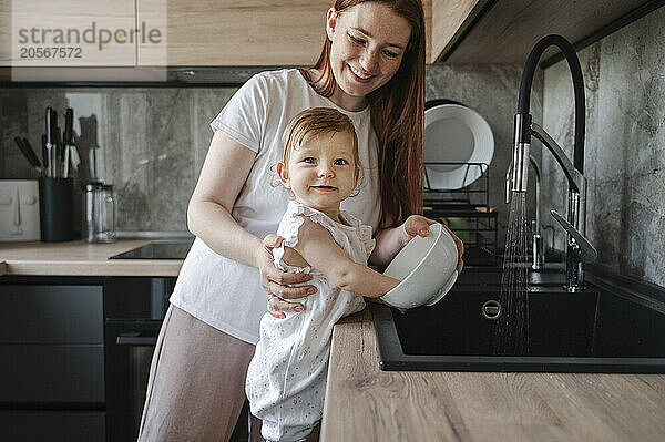 Smiling mother and daughter washing bowl in kitchen sink together at home