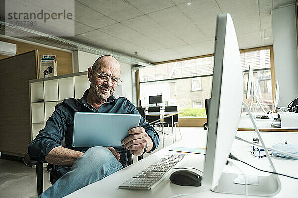 Mature businessman using tablet PC sitting at computer desk in office