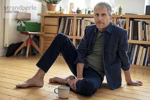 Retired senior man with coffee cup sitting on hardwood floor at home