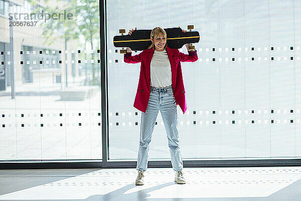 Smiling businesswoman with skateboard over shoulders standing at office