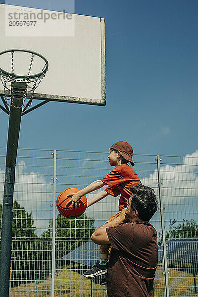 Boy throwing basketball in hoop and sitting on father's shoulder at court