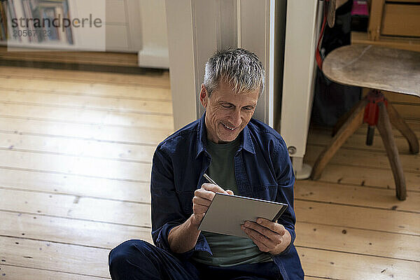 Smiling retired senior man writing in tablet PC sitting at home